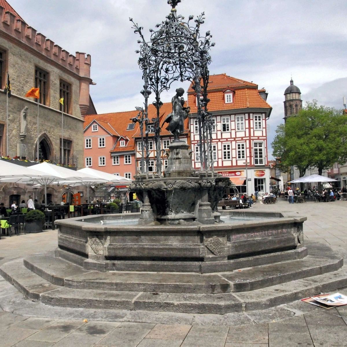 A wide shot of the Gänseliesel fountain in Göttingen, Germany, situated in a cobblestone town square. The central feature is an ornate, wrought-iron canopy structure covering a statue of a girl holding a goose. The fountain has a multi-tiered stone basin filled with water. In the background, there are traditional European buildings with red-tiled roofs, including a half-timbered house and a stone building with arched windows. People are seated at outdoor cafes under white umbrellas in the distance.