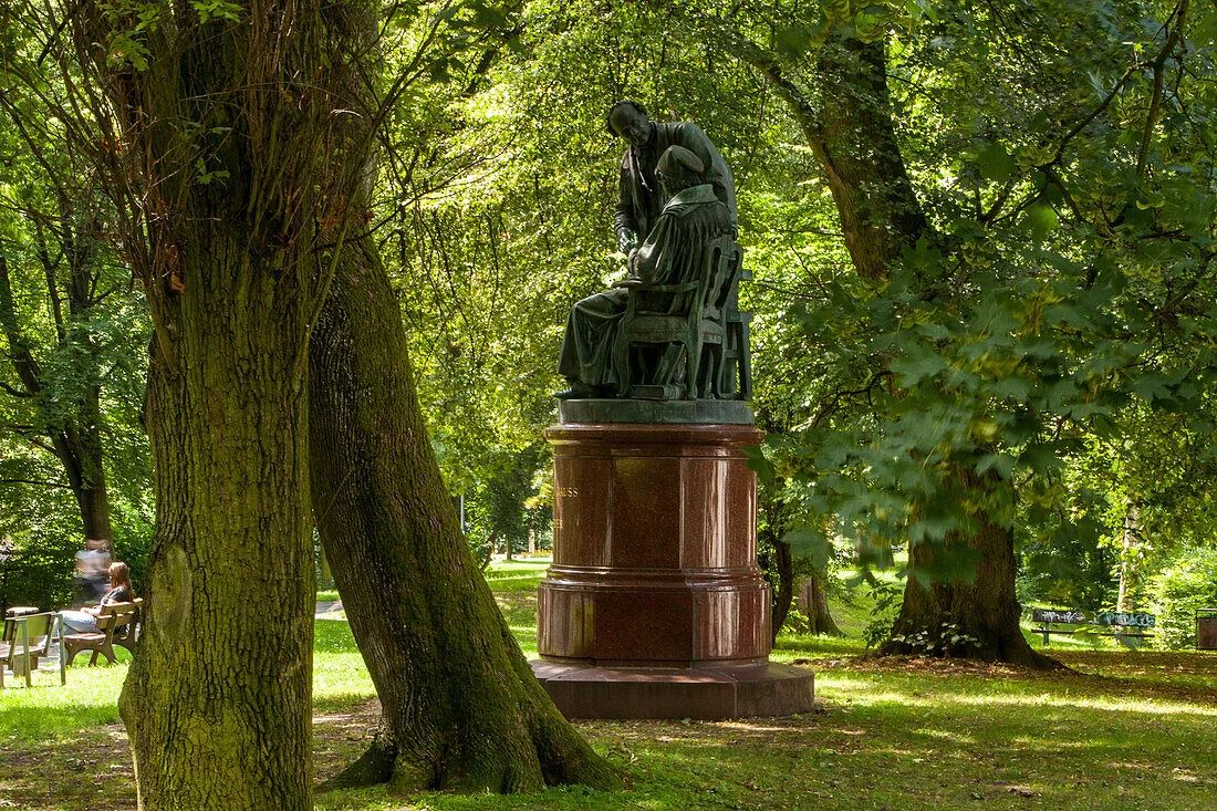 A bronze statue on a tall, reddish-brown stone pedestal situated in a lush green park. The statue depicts two figures: one seated man in robes looking down, and another standing man leaning over him, also looking down, as if in deep conversation or study. The statue is framed by large trees with thick trunks and green leaves, dappled with sunlight. In the background to the left, a person is sitting on a park bench.